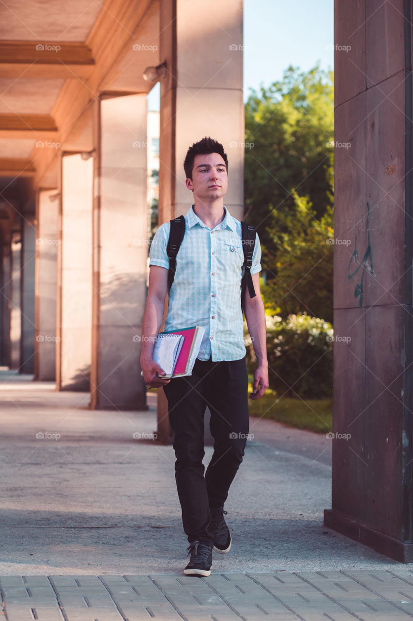 Student holding a notebook and carrying a backpack walking at the front of university building. Young boy wearing blue shirt and dark jeans