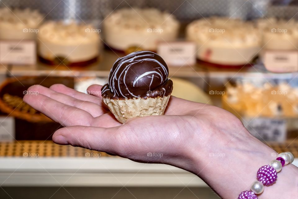 Small chocolate tart held on a hand on front of cakes and bakery goods