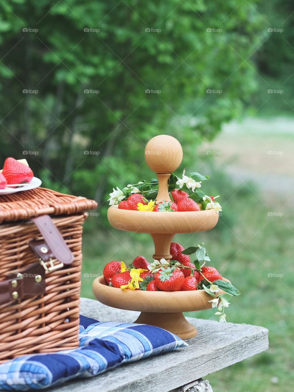 A bunk plate with ripe strawberries, spring flowers and a wicker basket stands on a bench in the park, close-up side view.