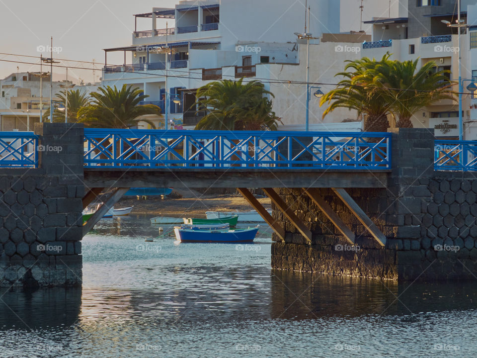 Paisaje de puente de piedra y madera en un entorno de pueblo de pescadores con palmeras y casas de fondo con la luz del atardecer.
