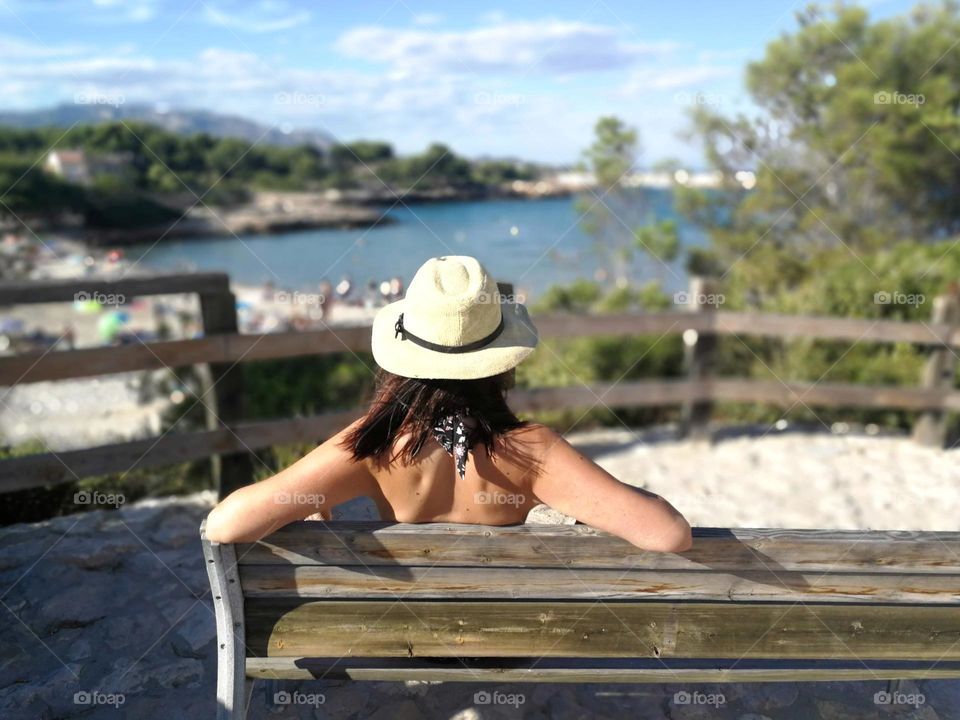 Summer time: in a sunny day, a woman with a hat is sitting on a bench in front of the sea