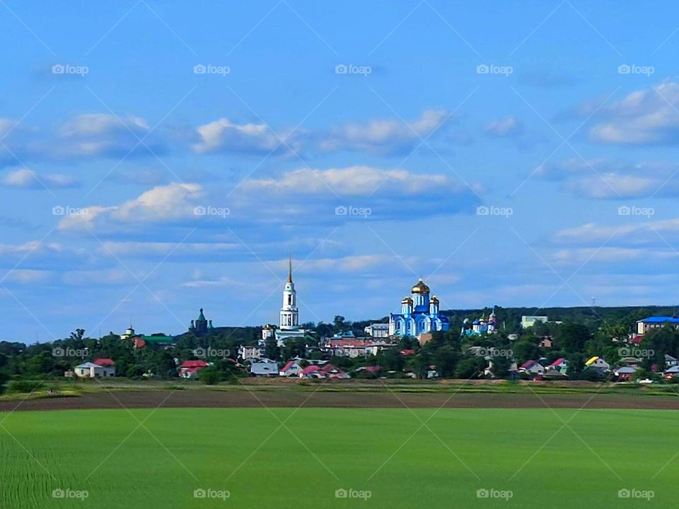 Countryside. Behind the green fields is a village in which the domes of the church rise. Blue sky with clouds enhances summer contrast