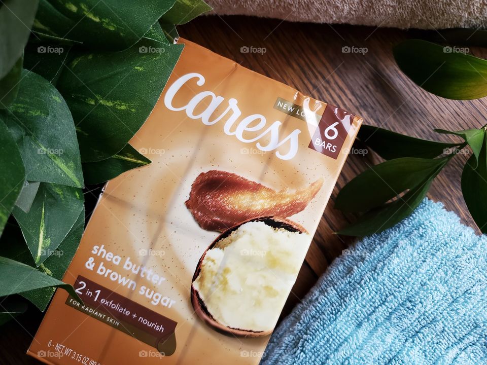 The new Caress bar six pack shea butter and brown sugar on a wood surface with a light blue towel,  a matching tan towel and greenery accents.