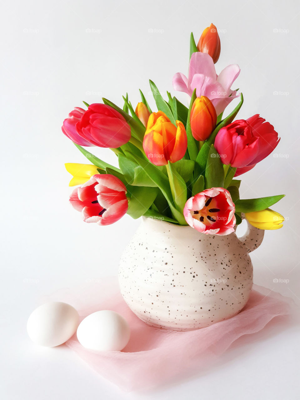 Spring holiday mood. Multicolored tulips in a jug with two whole raw white eggs on a white background. Preparing for Easter. Flower still life, vertical orientation