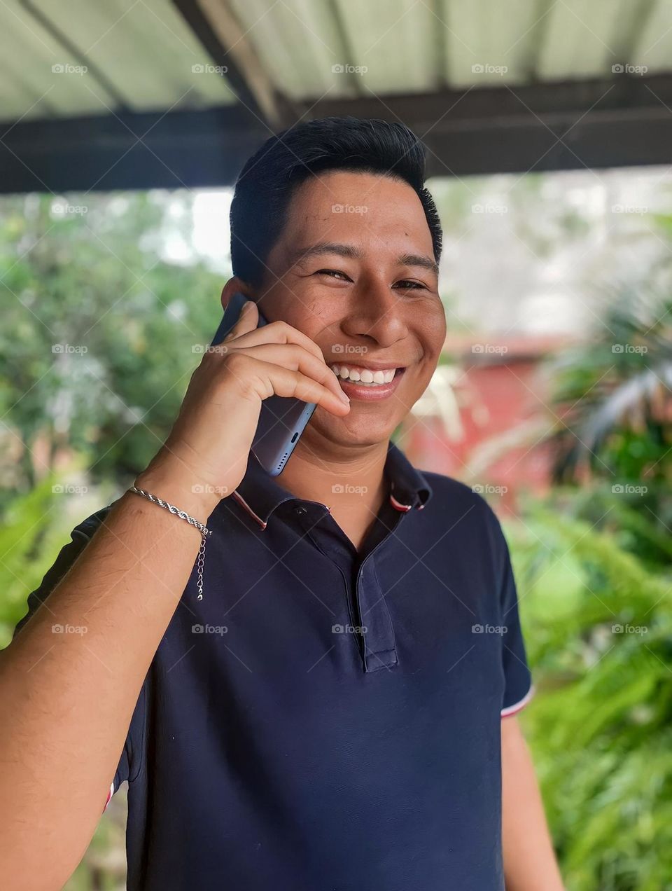 Young dark-skinned man smiling while talking on a light blue cell phone in a park with a garden