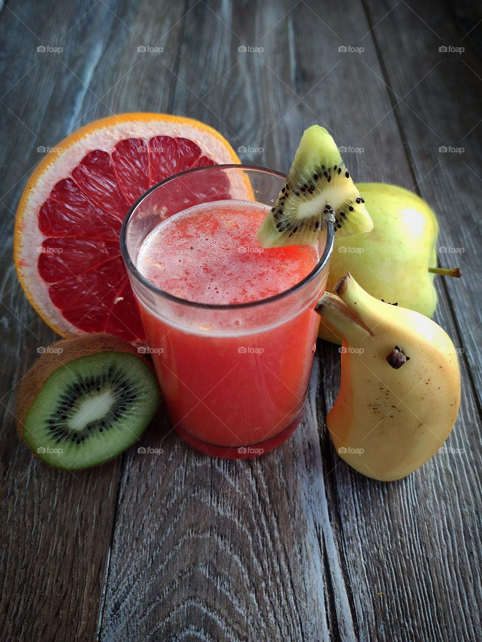 There is a glass with a red smoothie on a wooden surface.  There is a piece of kiwi on the glass.  Nearby are the ingredients: half a grapefruit, a green apple, half a kiwi, half a banana, which is decorated in the form of a dolphin