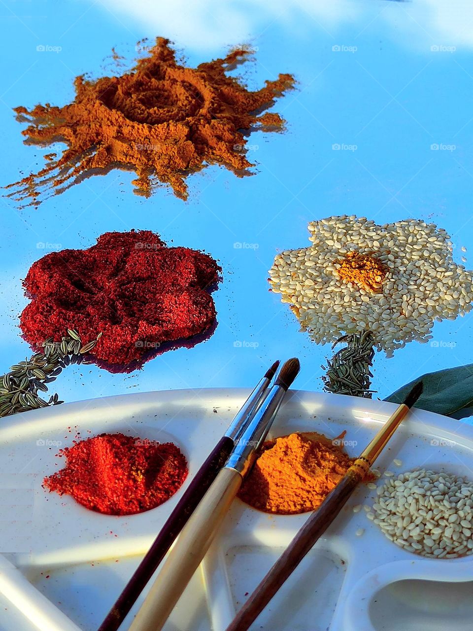 On the mirror surface of yellow curry sun,red turmeric flower with green fennel stem,white sesame chamomile and yellow curry.In the foreground lies a while palette with tassels, turmeric,curry and sesame seeds.Reflection of blue sky and white clouds