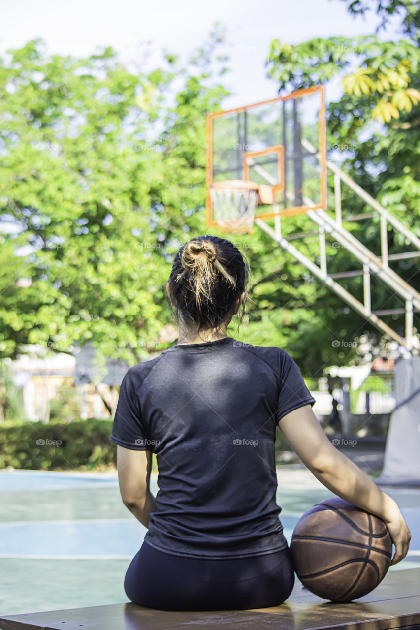 Asian Woman holding basketball sitting on a wooden chair Background blur basketball hoop and tree in park.