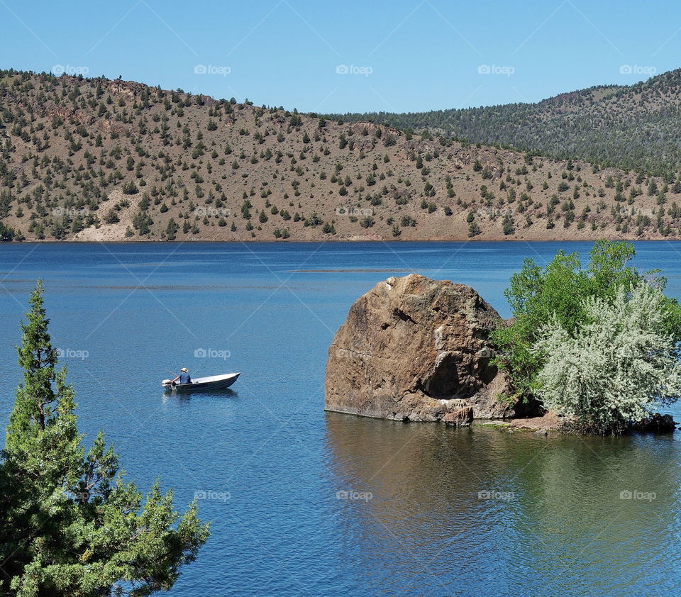 A fisherman in a boat trolling by a large boulder in Prineville Reservoir in Central Oregon on a sunny summer morning. 