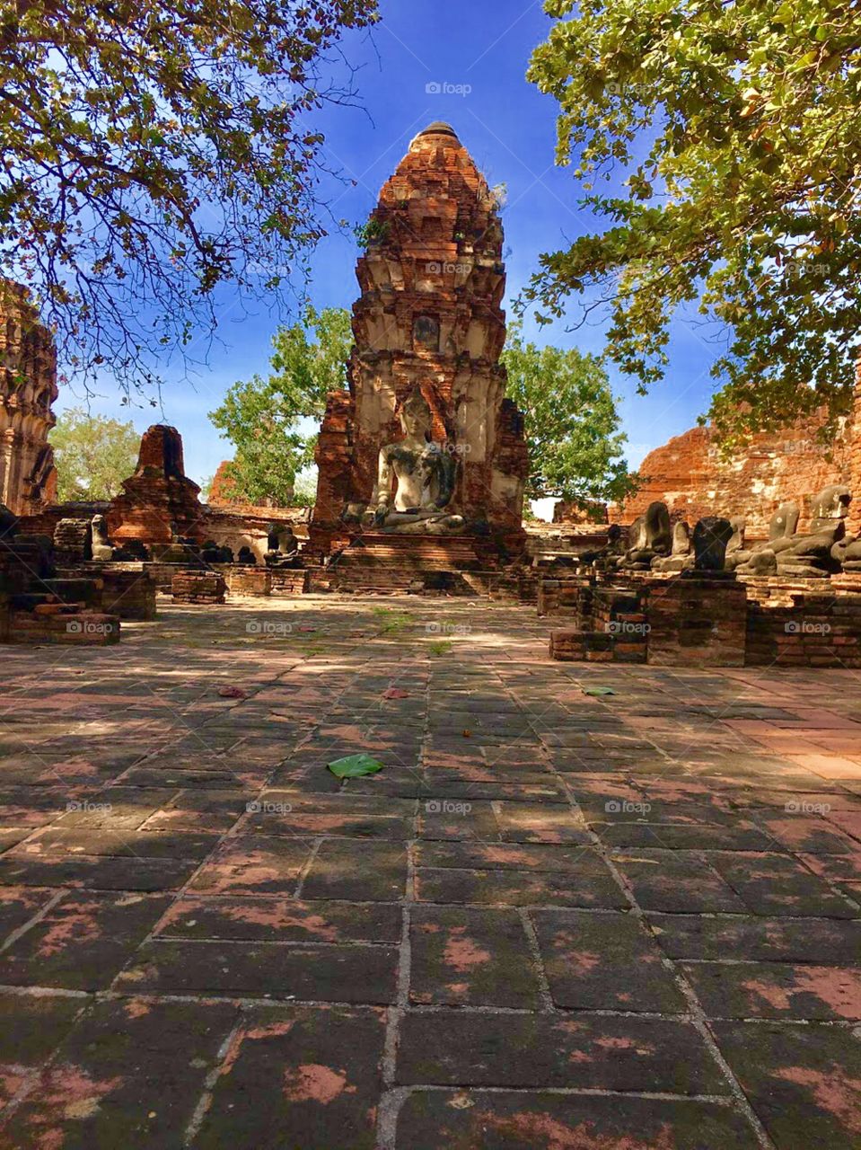 Temple ruins in Ayutthaya in front of the blue sky surrounding by green trees as the reddish colors of the bricks  stand out in different shades.