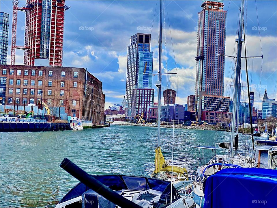 A closeup of the picturesque & versatile “Newtown Creek” with its many different kinds of boats, spectacular views of the Manhattan as well as the LIC, Queens skyline. Across the East River a tall building is in the works. 2022. Hypnotic Productions