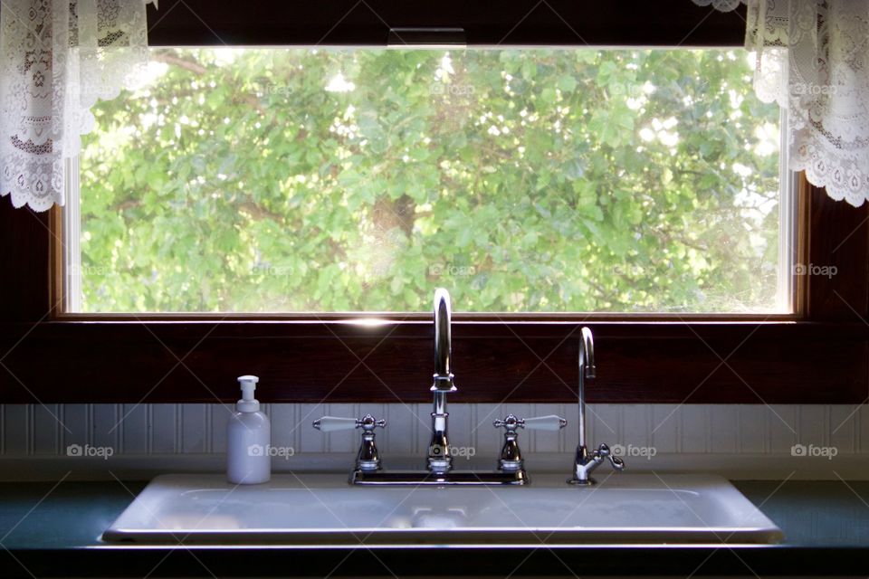 A white kitchen sink with vintage-style chrome fixtures by an open window, soft sunlight through a large leafy tree visible