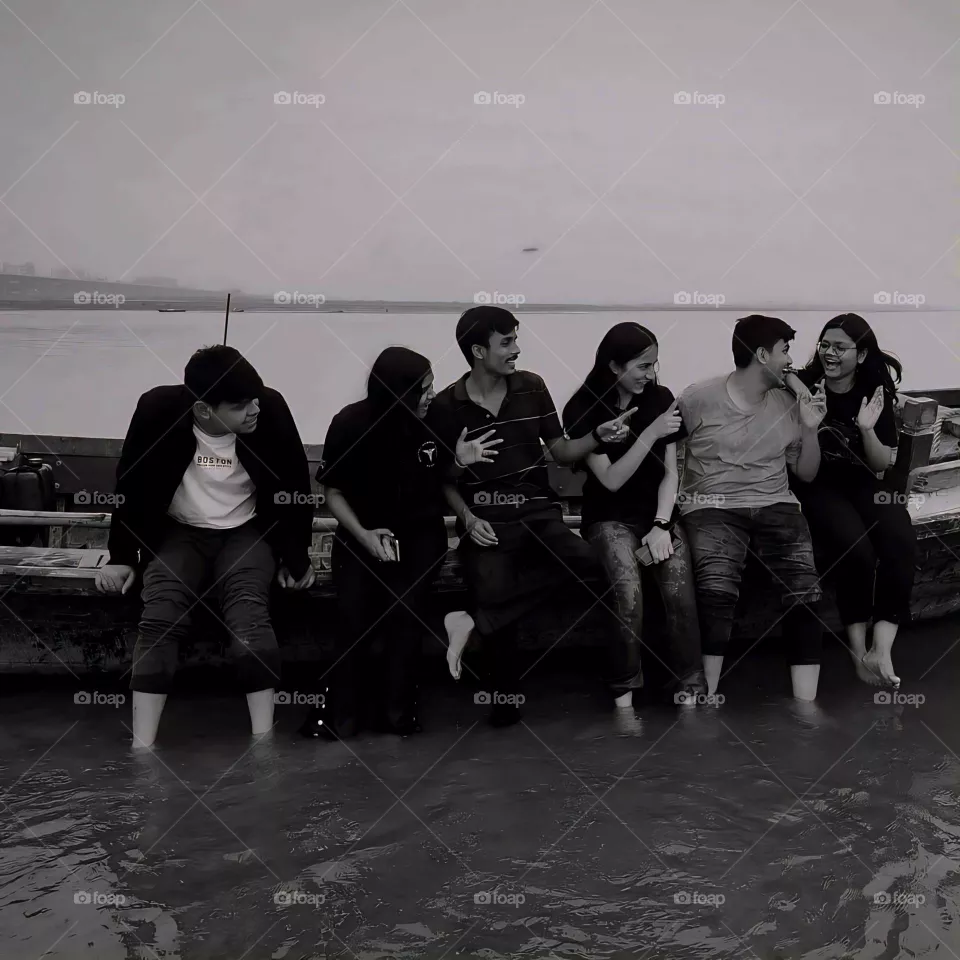The monochrome palette adds a timeless quality, highlighting the friends' emotions and expressions. Their casual, barefoot postures emphasize relaxation and spontaneity, capturing a beautiful, carefree moment in a tranquil setting.