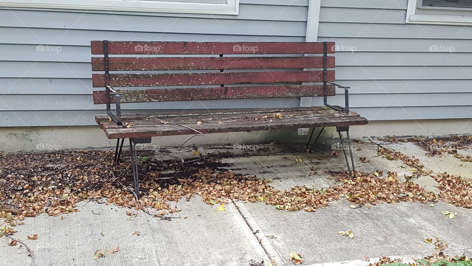 A worn out red bench sitting in front of our house. The other part of the bench is on our back porch and together they make a picnic table. There are leaves everywhere on the ground.