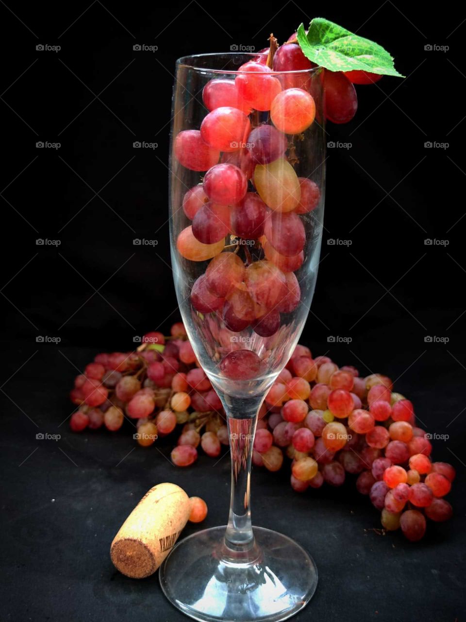 A wine glass containing a red bunch of grapes. Next to the glass is a red bunch of grapes and a wooden cork from a wine bottle. Black background