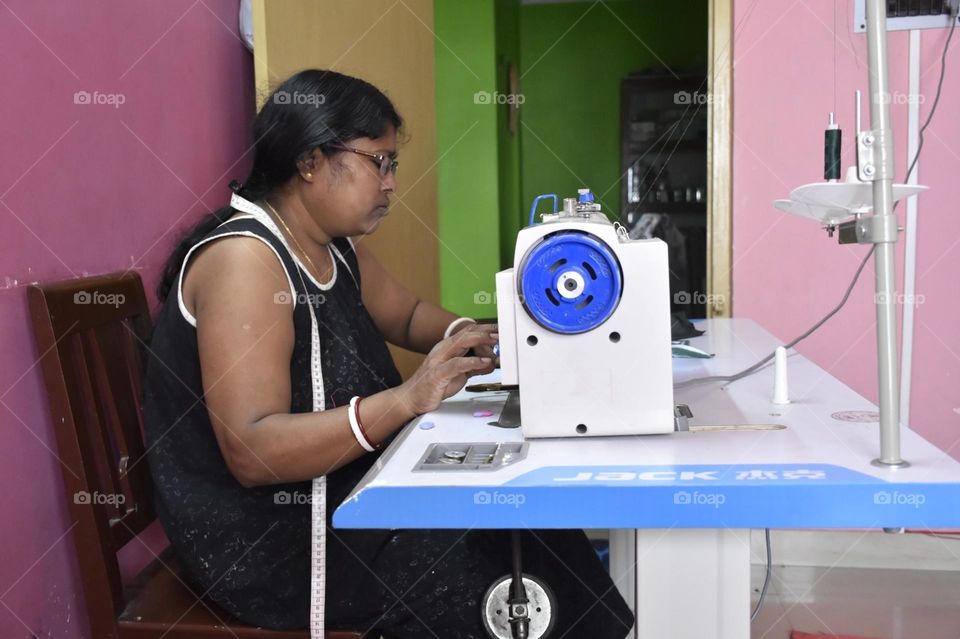 One woman is engaged with her sewing machine. Tailoring is her profession. She is making a garment with her sewing machine and she keeps a tailoring tape in her shoulder. This photo of women and how they are contributing to our society.