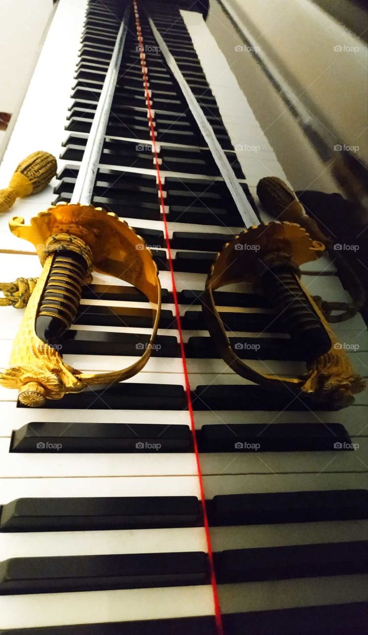 Reflection from the surface of the piano. On the keys of the piano lies the sword of an Italian air force officer. In the foreground there is a golden sword hilt, which turns into a metal blade. The sword is reflected from the black polished surfa