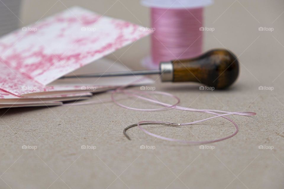 On a sand-colored background are tools for manual binding a needle, pink thread, small papers with pink patterns