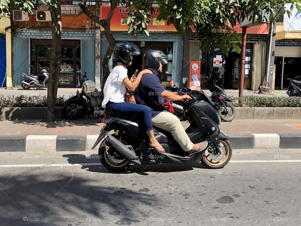 Daily life motorbike travel. Asian couple with helmets. Daytime. In motion. Bali, Indonesia. 2018.
