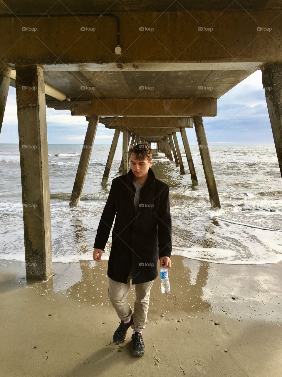 Stylish young man wearing black wool coat and holding water bottle walking toward camera under ocean Jetty 