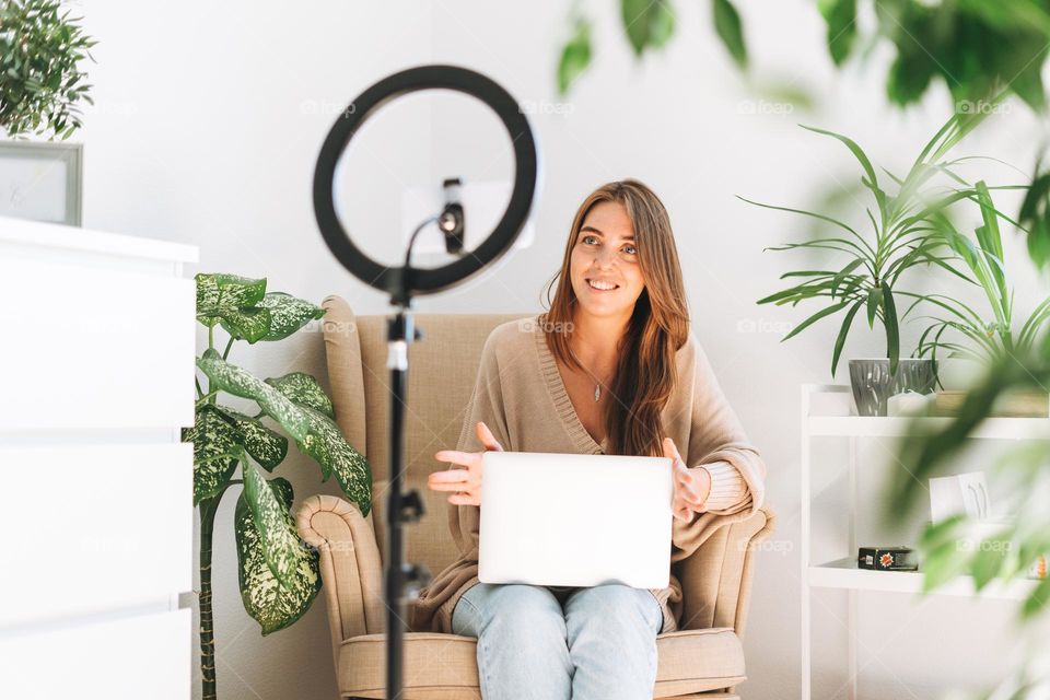 Young pretty woman blogger records video on mobile phone using ring lamp sitting on chair with laptop in room with green plants at home