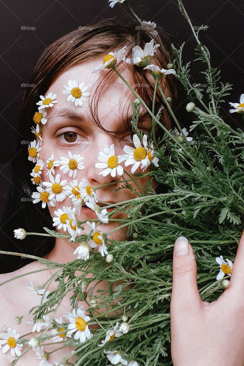 conceptual, original portrait of a girl with a bouquet of field daisies and chamomile buds on half of her face with brown eyes