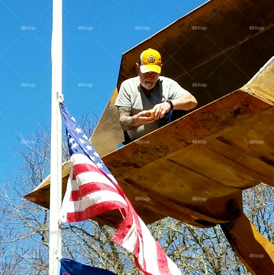 Man in bucket front end loader, up position. He's fixing flagpole with lowered flag blowing in wind.🇺🇸Sunny day, cap on worker. Hands fixing tools.