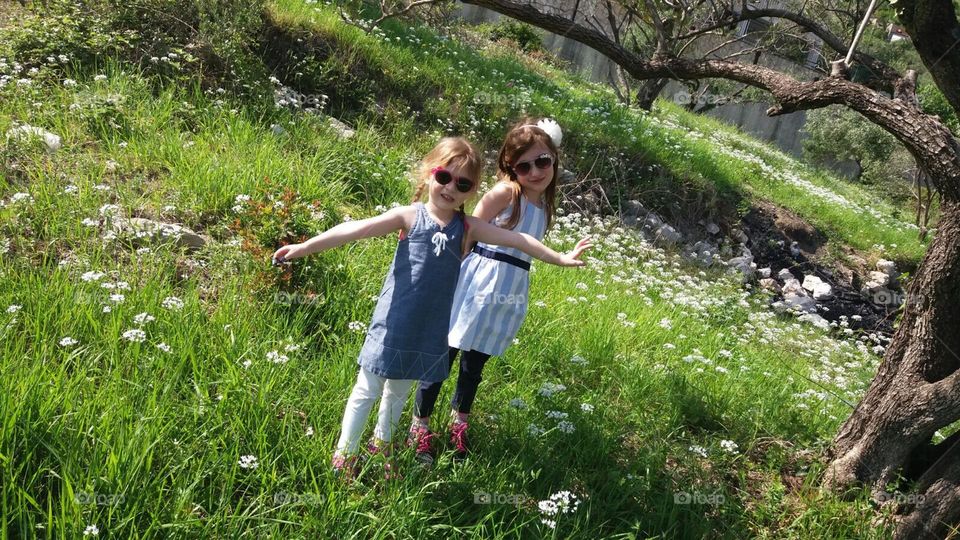 Two young girls in springtime dresses picking wildflowers near an olive tree on a warm spring day.