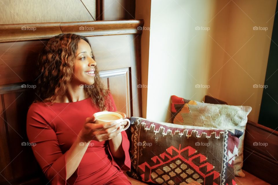 Diverse woman in red happily holds coffee in cozy corner. She has curly hair and looks towards the window light. 