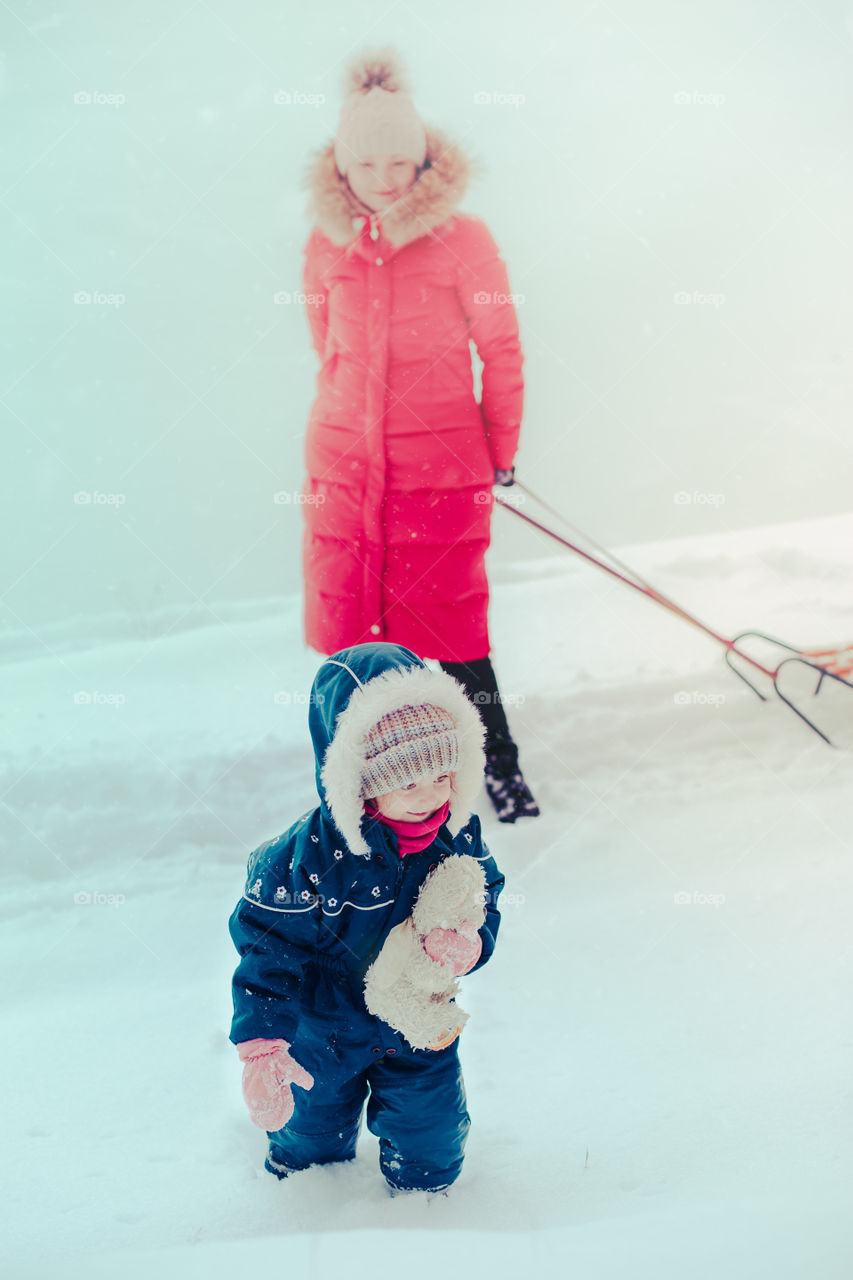 Mother and her little daughter are spending time together walking outdoors in winter while snow falling. Woman is pulling sled, a few years old girl is walking through the deep snow, enjoying wintertime