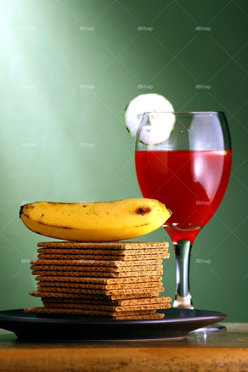 stack or pile of soda crackers, a banana and a goblet of fruit juice with cucumber slices