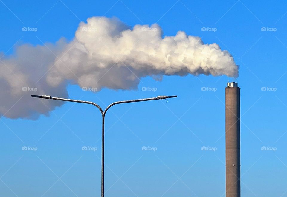 Minimalistic view with white grey smoke from the high tall production pipe and the streetlight against the bright blue sky