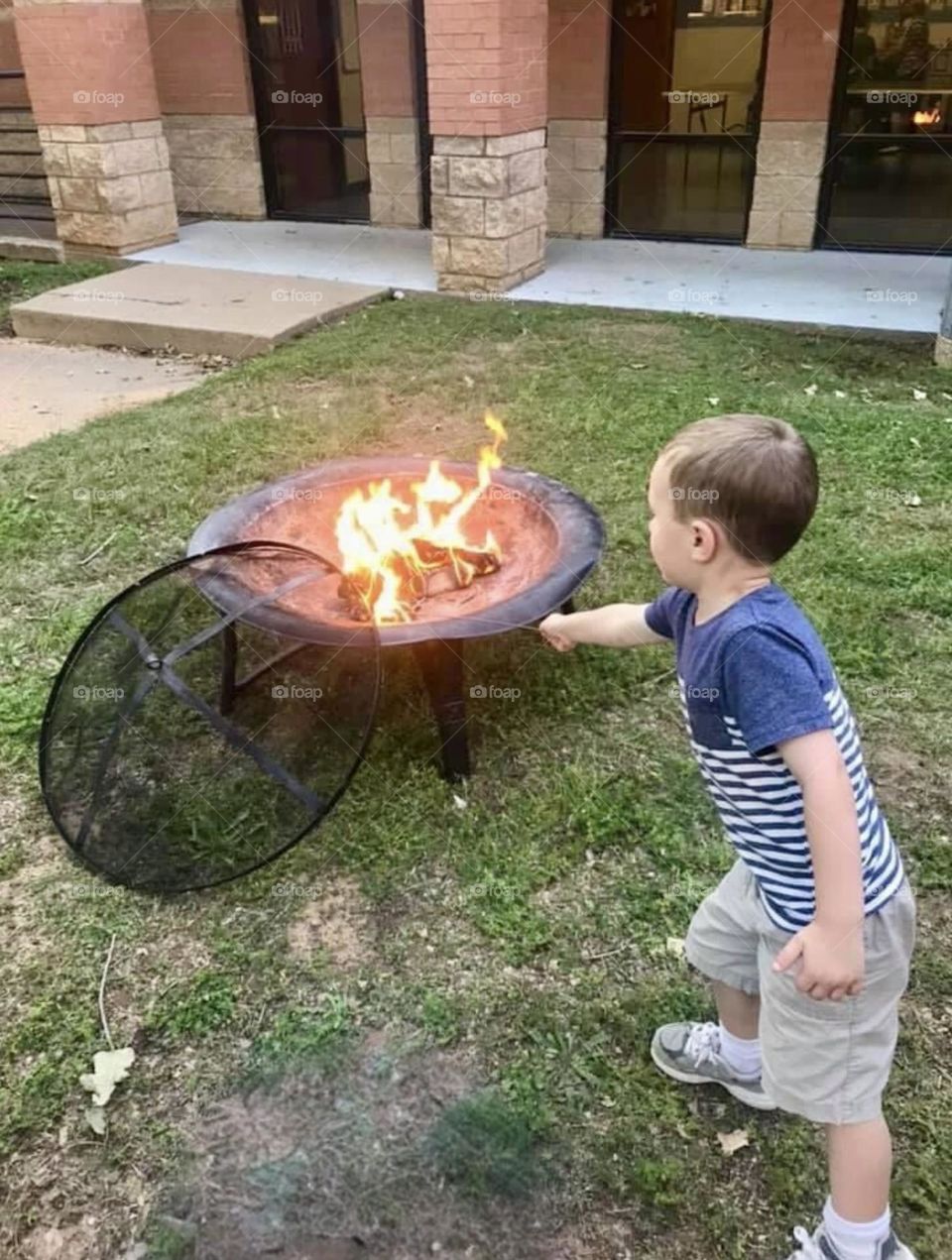 A young boy roasts a marshmallow in a campfire outside over the green grass. 