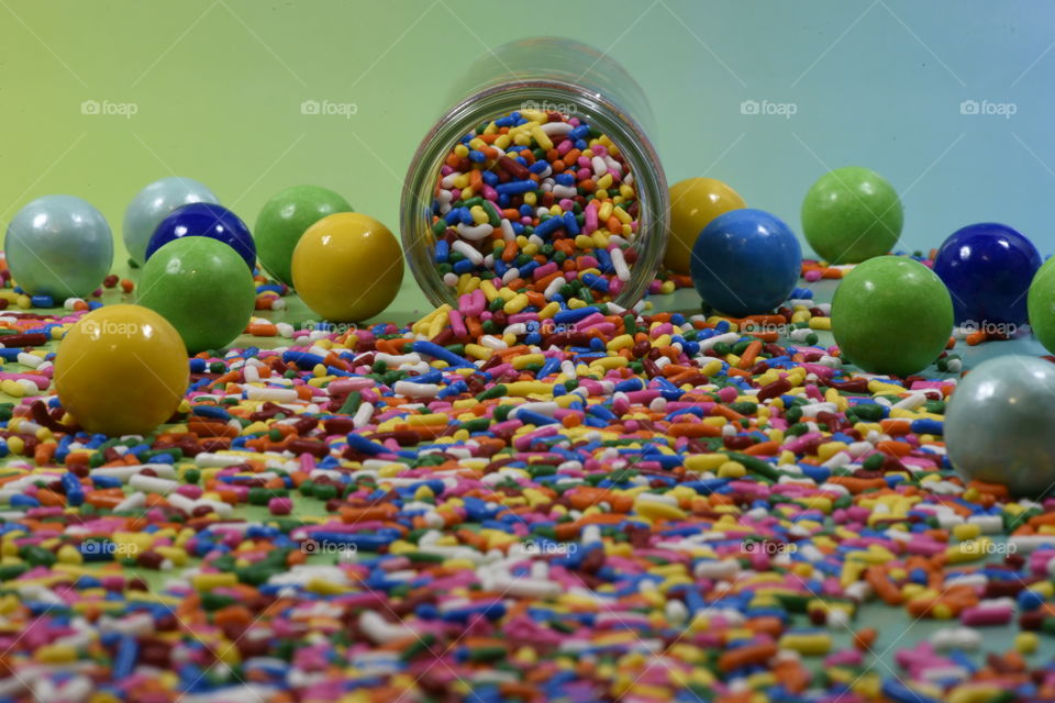 Multicolored sugarcoated gumballs and sprinkles spill out of a jar
