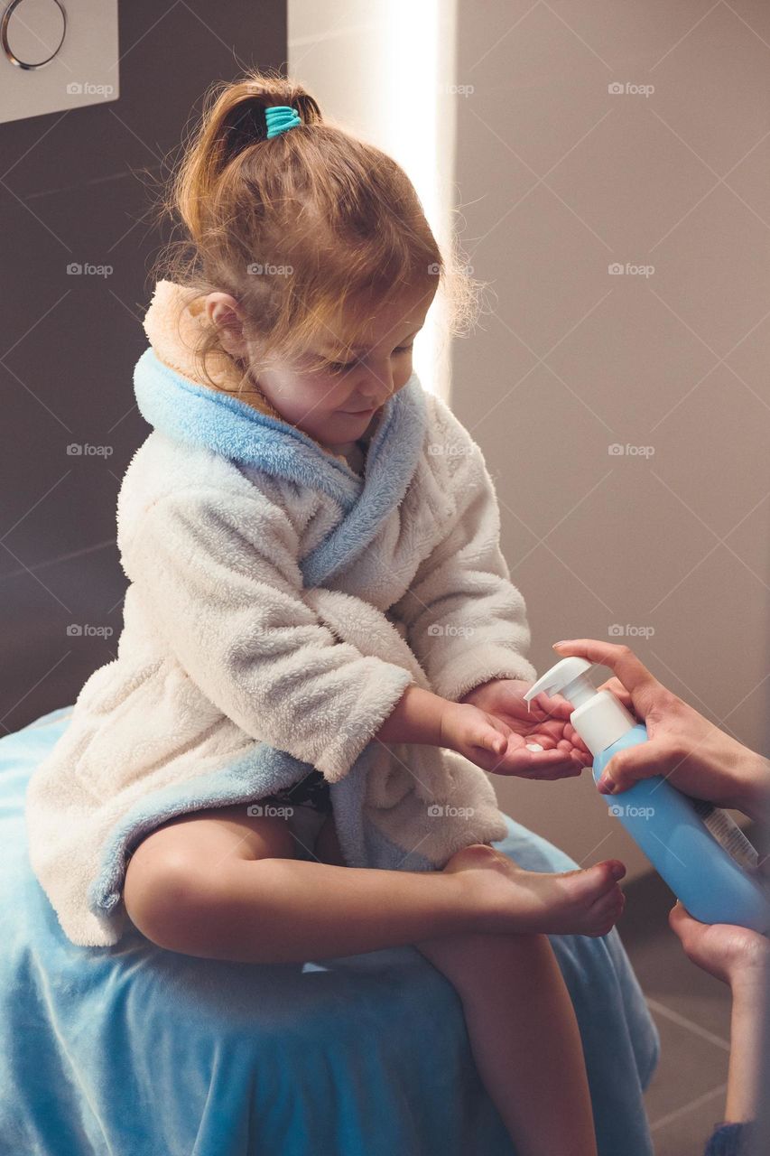 Mother applying moisturizing cream on her daughter's legs after bath. Mom caring about her child. Girl sitting in bathroom, wearing bathrobe