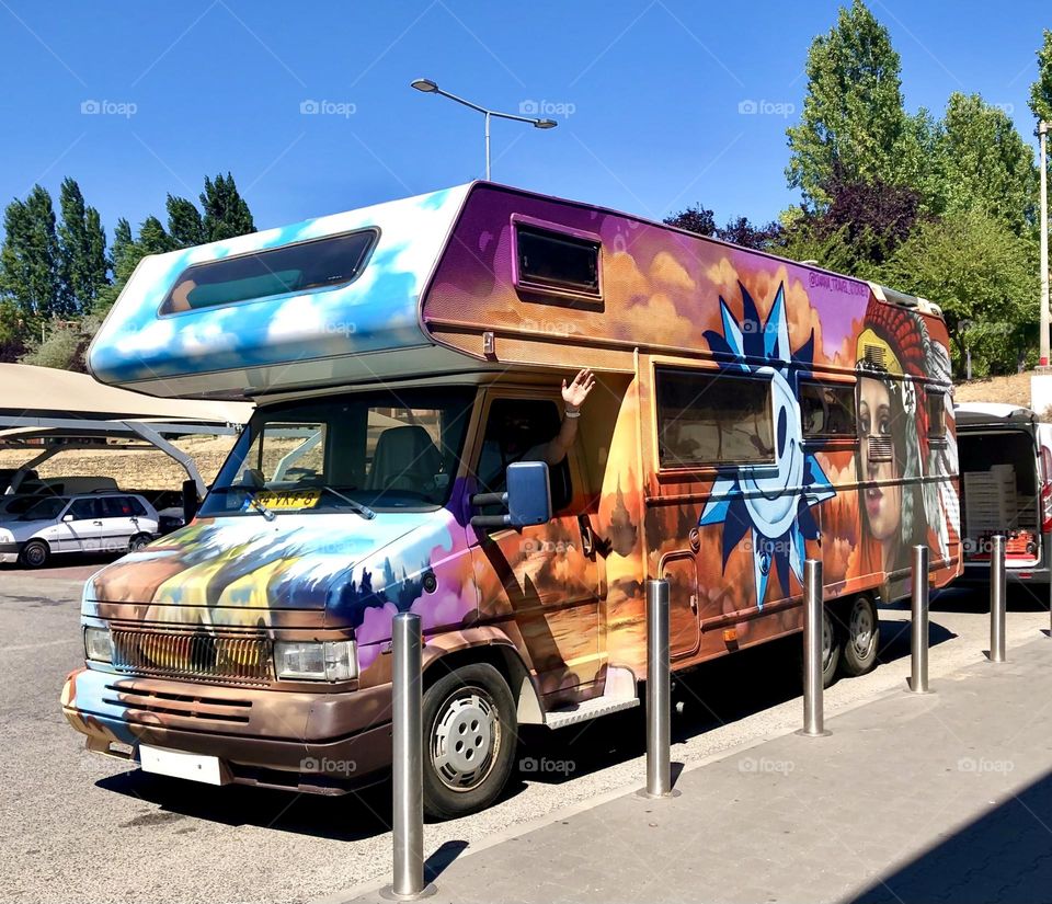 Brightly coloured camper van covered with murals and a friendly waving hand coming out the window