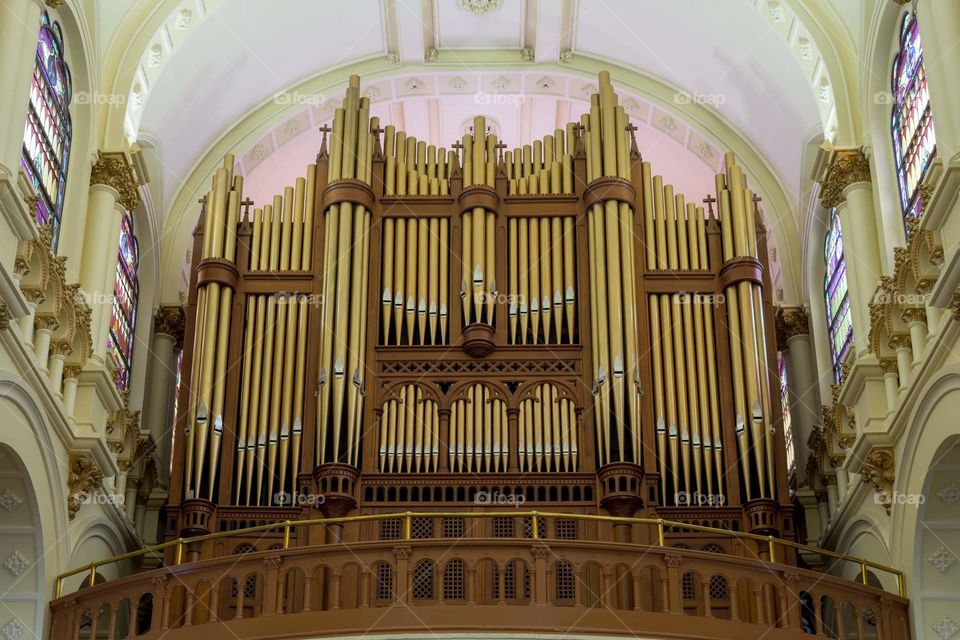 pipe organ in cathedral with gold pipes and wood frames and railings as a pink hue fills the ceiling from the light through the stained glass