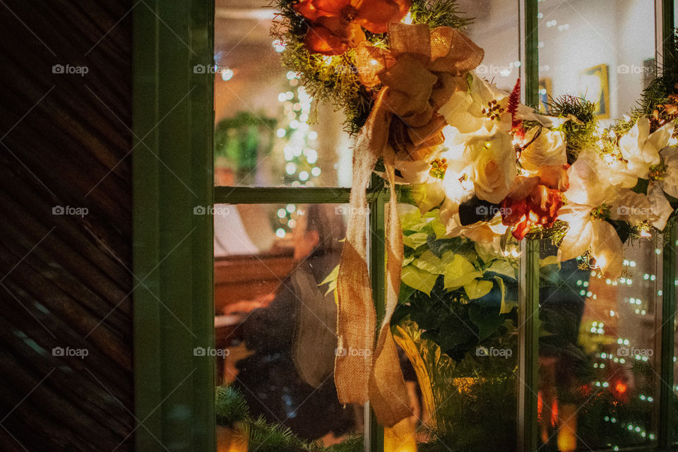 Christmas wreath on the house window with an old lady playing piano/Christmas carols seen through the glass window.