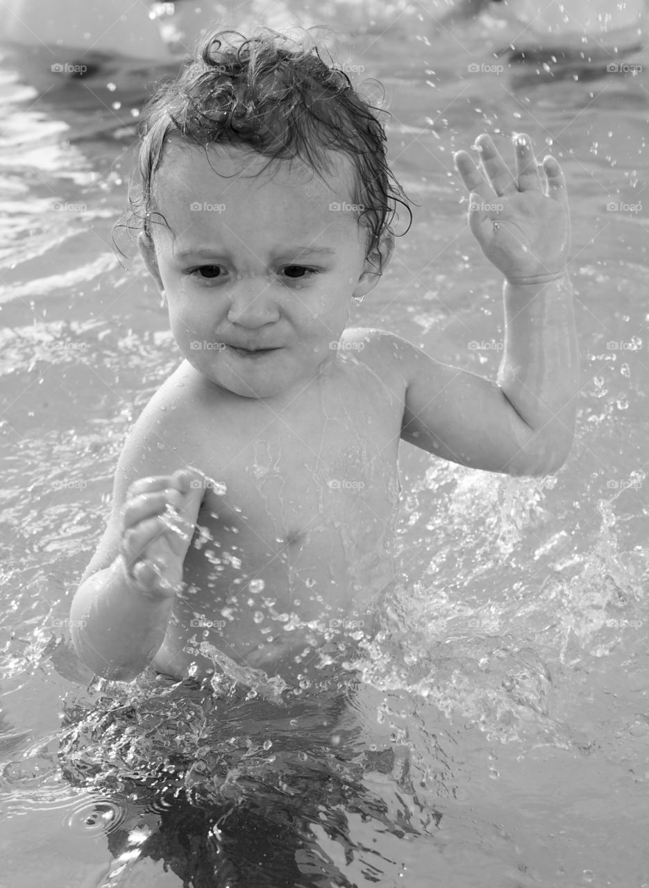 A toddler boy plays and splashes in an outdoor swimming pool on a sunny summer day. 