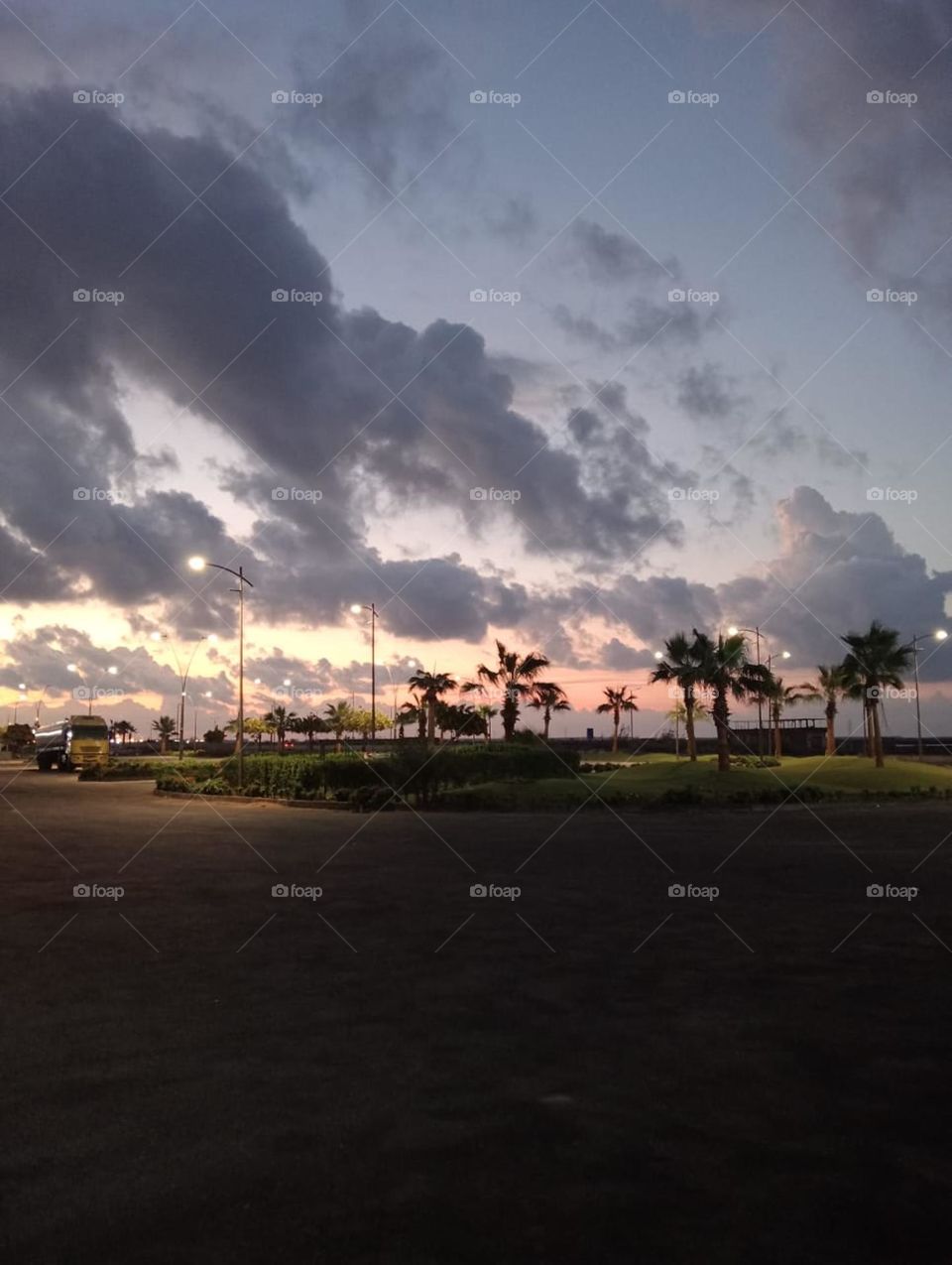 a sunset scene in a coastal area or a public park near the sea. The sky is filled with dark clouds that contrast beautifully with the warm sunset colors, displaying a mix of blue, gray, and soft orange near the horizon,