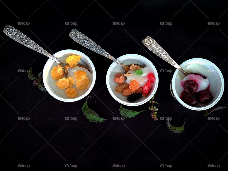 Food. On a black background are three jars of white pudding. On the surface of each pudding are different ingredients: nut mix, corn flakes and frozen cherries. Each jar contains a metal spoon. Green basil leaves all around