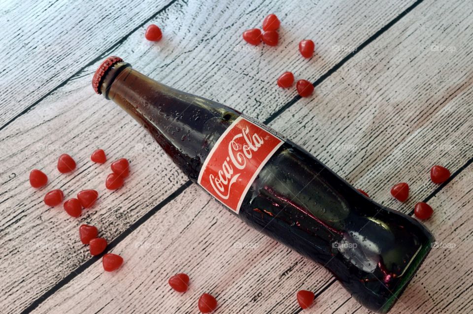 A glass bottle of Coca-Cola with candy hearts on a white rustic background