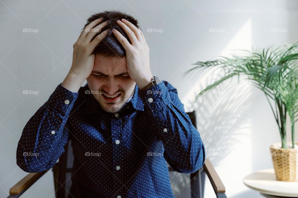 Portrait of a beautiful Caucasian young guy in a shirt with disappointed emotions holding his head with both hands standing in a chair in a room on a summer day, side view close-up.