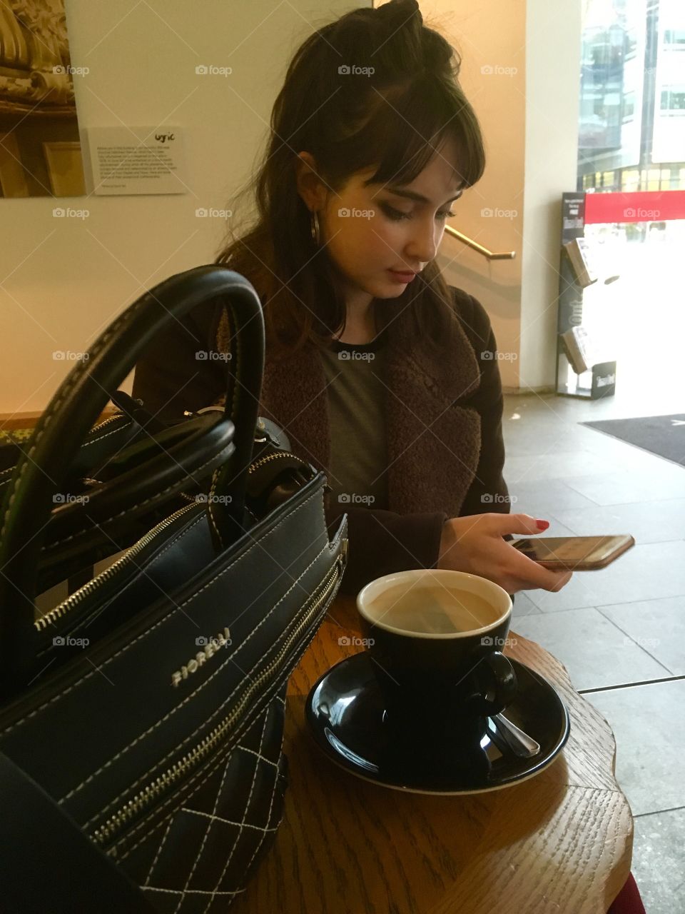 Pretty young woman, coat and bag. Sitting at table using her phone