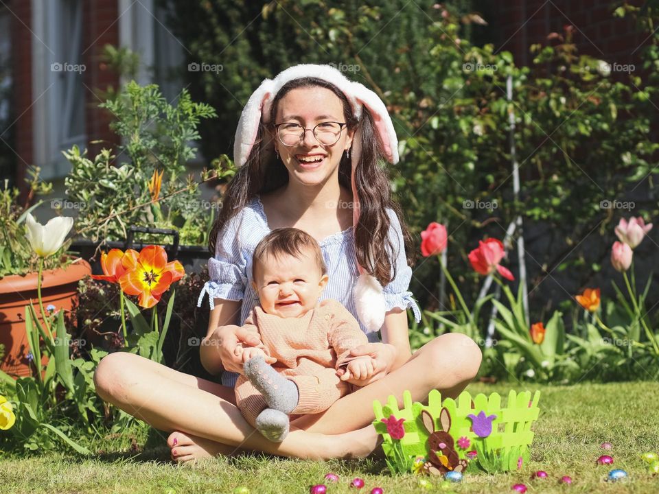 Two beautiful and happy caucasian girls sisters are sitting on the lawn in the backyard of the house with a felt basket and chocolate eggs, celebrating Easter according to tradition on a sunny spring day, close-up side view. Concept happy kids, celeb