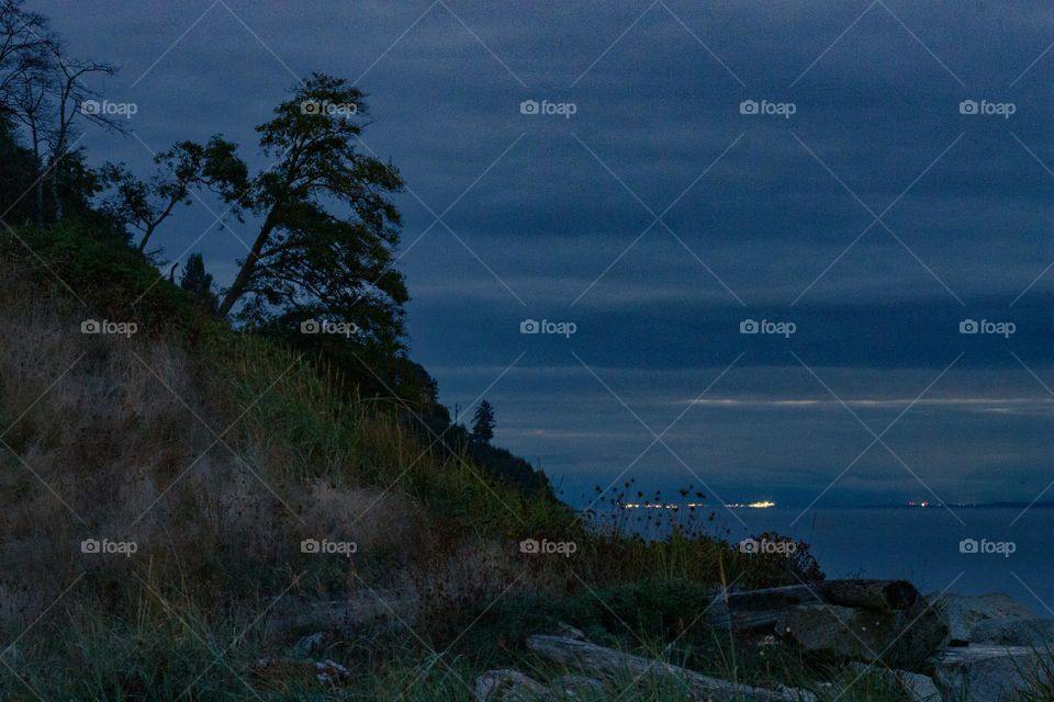 Twilight shot of the beach cliff and the strait. The lights of a town on the other side of the strait add some light and depth to the darkening vista. 