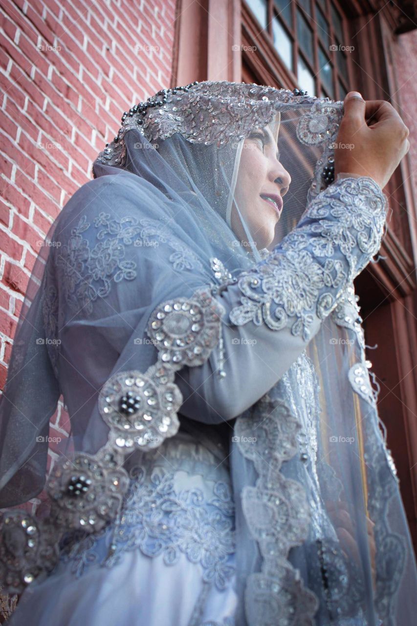 Portrait of young and beautiful Asian muslim woman wearing silver robe with matching hijab and headscarf, holding headscarf and smiling, Red brick wall background. for the wedding.