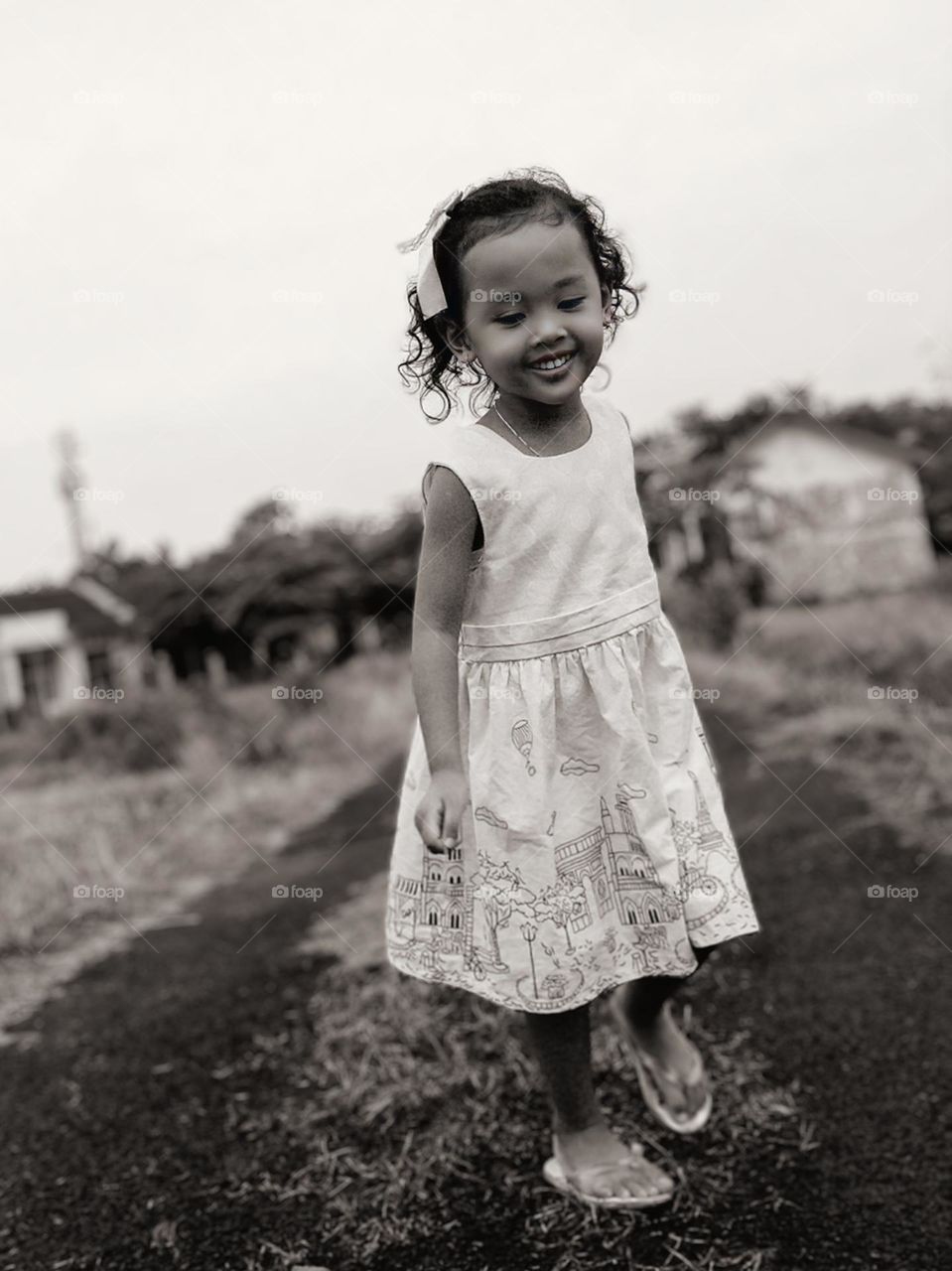 Close-up of a child with a cheerful face, wearing a short-sleeved dress with a building motif, standing outdoors. The child is standing on a dirt road or path