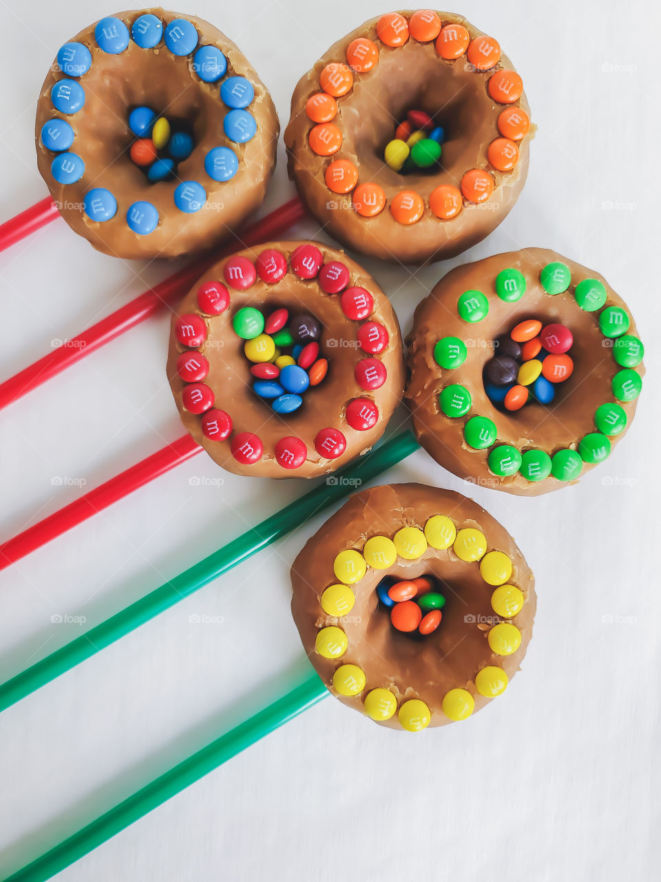 M&M minis decorated donuts filled with peanut M&Ms and mini M&Ms in the center, layed out in a creative bursting/ shooting star or flower bouquet design on a white background. Open to creative interpretation.