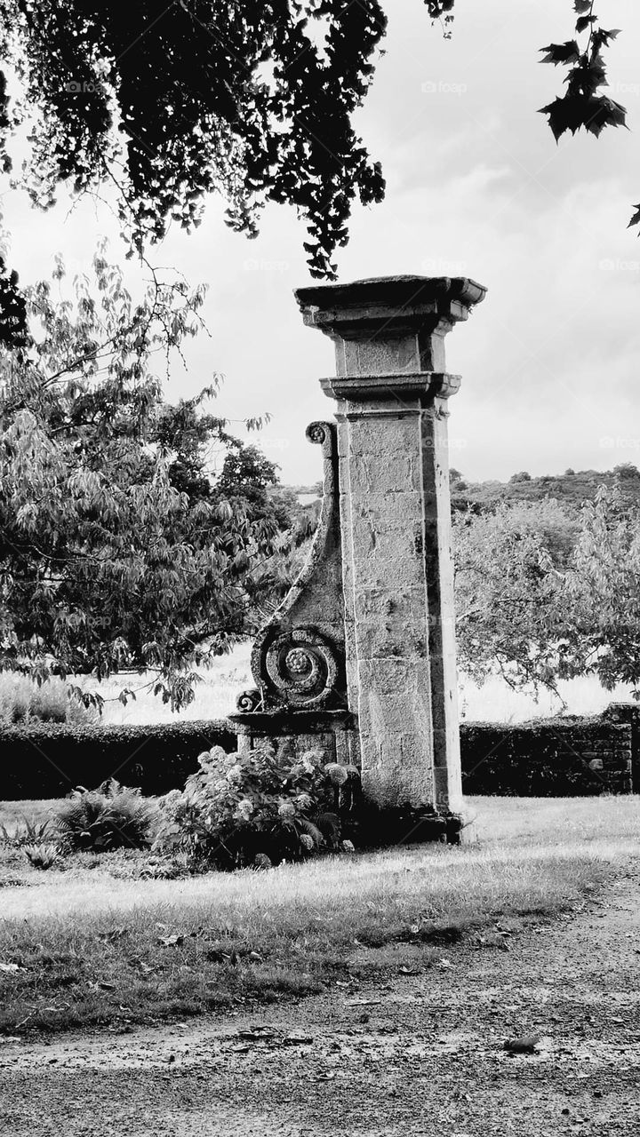 Black and white shot of a vintage looking 13th century stone gate pillar standing alone on grass and among hydrangea, shrubs and trees, only remaining part of the entrance gate of Beauport's abbey